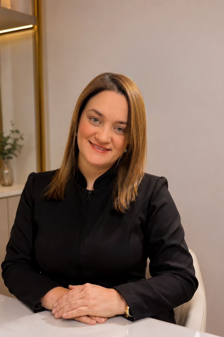 Keyla Miranda de jaqueta preta, sentada à mesa branca, com as mãos cruzadas.